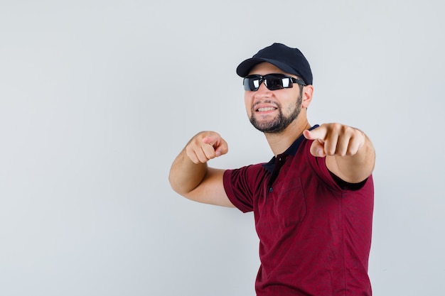 Young male in t-shirt,sunglasses pointing at camera and looking cheery , front view.