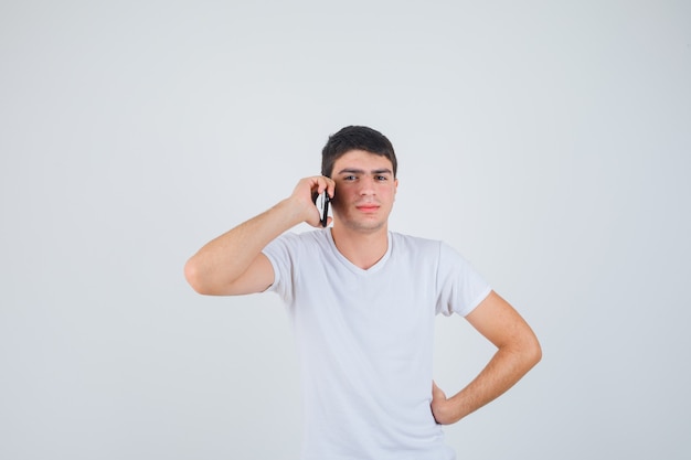 Молодой мужчина в футболке разговаривает по мобильному телефону и выглядит уверенно, вид спереди.