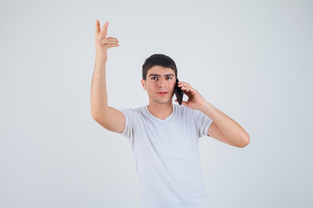 Young male in t-shirt talking on mobile phone while raising arm and looking focused , front view.