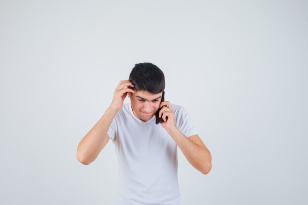 Young male in t-shirt talking on mobile phone while scratching head and looking thoughtful , front view.