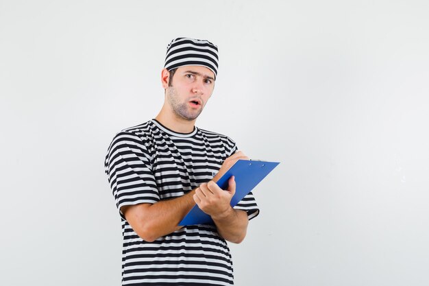 Young male taking notes on clipboard in t-shirt, hat and looking hesitant , front view.