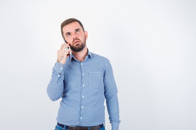 Young male talking on mobile phone in shirt, jeans and looking thoughtful. front view.