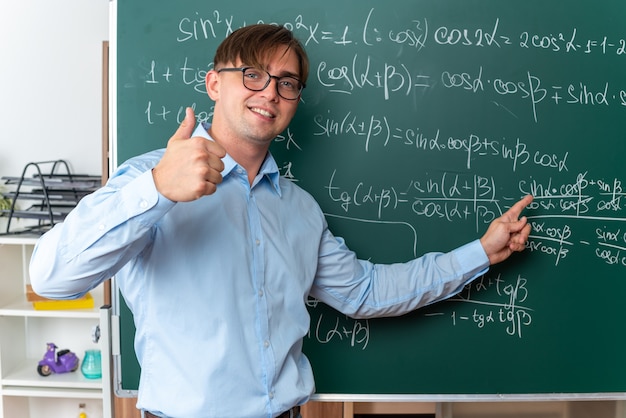 教室で数式を使って黒板の近くに立って親指を立ててレッスンを説明する眼鏡をかけた若い男性教師