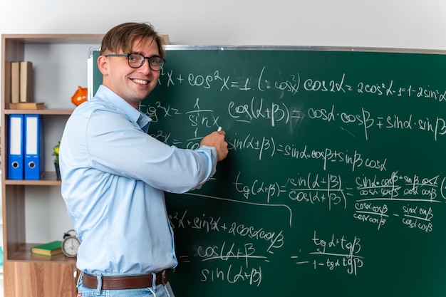Foto gratuita giovane insegnante maschio con gli occhiali che spiega la lezione felice e sorridente in piedi vicino alla lavagna con formule matematiche in classe