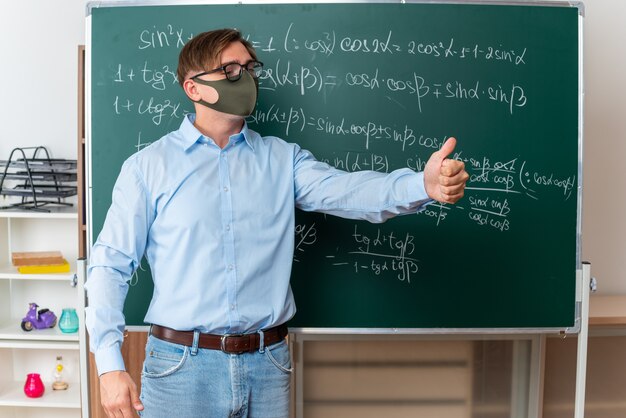 Young male teacher wearing glasses in facial protective mask explaining lesson showing thumbs up standing near blackboard with mathematical formulas in classroom