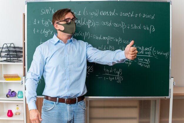 Молодой учитель-мужчина в очках в защитной маске для лица объясняет урок, показывая большие пальцы руки вверх, стоя возле доски с математическими формулами в классе