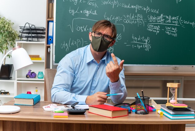 Young male teacher wearing glasses and facial protective mask looking confused raising arm in displeasure sitting at school desk with books and notes in front of blackboard in classroom