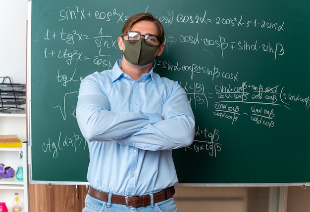 教室で腕を組んで自信を持って見える数式を使って黒板の近くに立つ顔面保護マスクに眼鏡をかけた若い男性教師