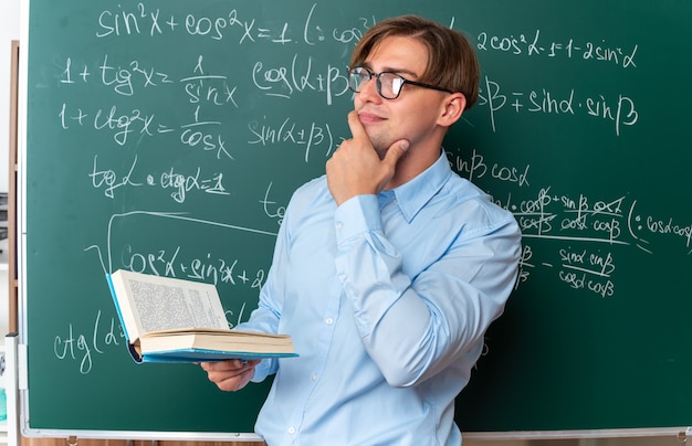 Young male teacher wearing glasses holding book looking aside with hand on chin thinking standing near blackboard with mathematical formulas in classroom