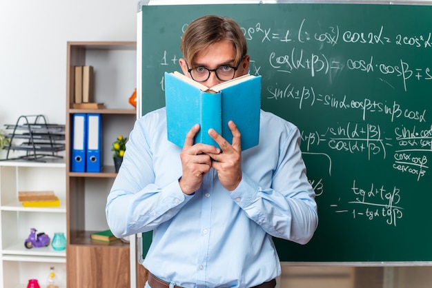 Free photo young male teacher wearing glasses holding book  with serious face standing near blackboard with mathematical formulas in classroom