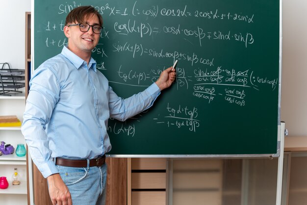 교실에서 수학 공식으로 칠판 근처 자신감 서 미소 수업을 설명하는 분필을 들고 안경을 착용하는 젊은 남성 교사