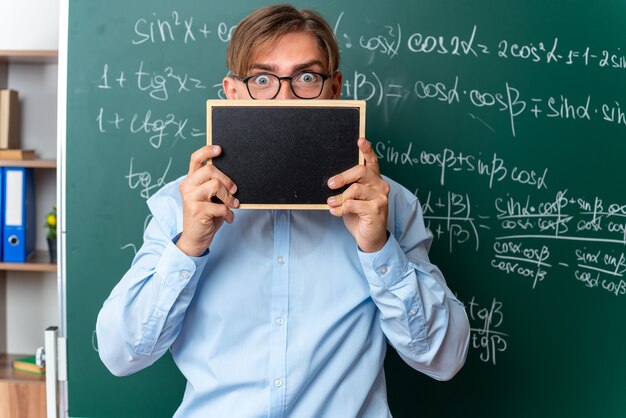 Young male teacher wearing glasses holding small blackboard in front of his face looking surprised standing near blackboard with mathematical formulas in classroom