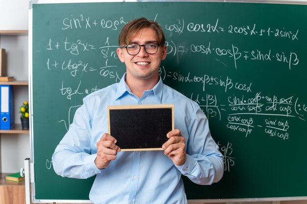 Young male teacher wearing glasses holding small blackboard looking smiling confident standing near blackboard with mathematical formulas in classroom