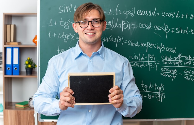 Молодой учитель-мужчина в очках держит небольшую доску, улыбаясь, уверенно стоит возле доски с математическими формулами в классе