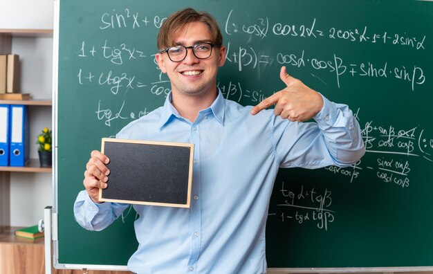 Молодой учитель-мужчина в очках держит небольшую доску, указывая на нее указательным пальцем, уверенно улыбаясь, стоя возле доски с математическими формулами в классе