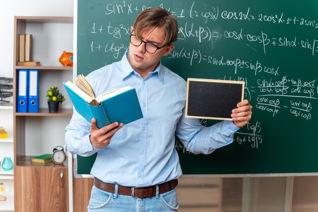 Young male teacher wearing glasses holding small blackboard reading book looking confident standing near blackboard with mathematical formulas in classroom