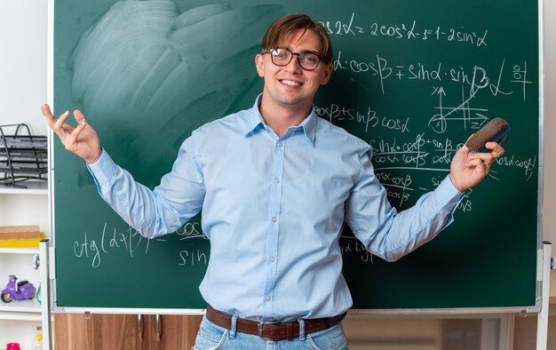 教室で数式を使って黒板の近くに自信を持って立って笑顔のスポンジを持つ眼鏡をかけた若い男性教師