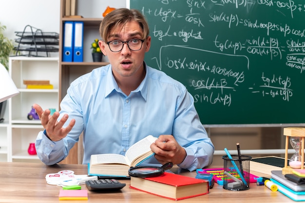깜짝 놀라게 찾고 안경을 쓰고 젊은 남성 교사는 교실에서 칠판 앞에 책과 메모와 함께 학교 책상에 앉아