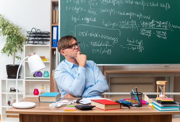 Young male teacher wearing glasses looking aside with hand on chin with pensive expression thinking sitting at school desk with books and notes in front of blackboard in classroom