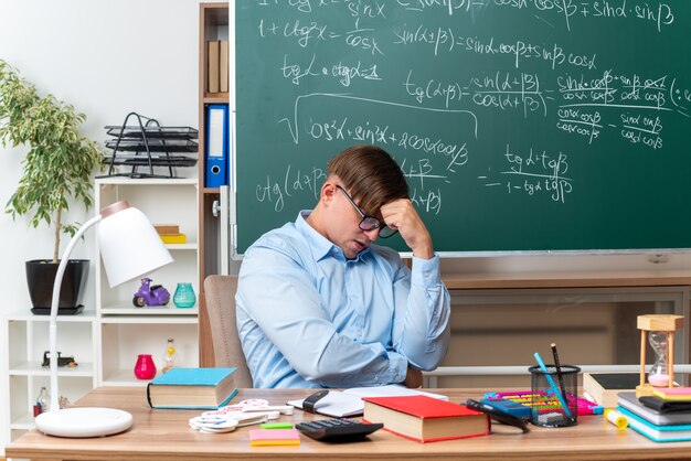 교실에서 칠판 앞에 책과 메모와 함께 학교 책상에 앉아 혼란과 매우 불안 찾고 안경을 착용하는 젊은 남성 교사