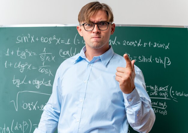 Young male teacher wearing glasses looking displeased and angry standing near blackboard with mathematical formulas in classroom