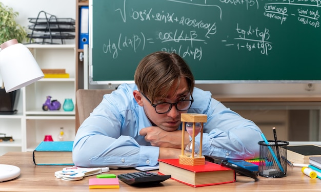 Young male teacher wearing glasses looking at hourglass tired and bored sitting at school desk with books and notes in front of blackboard in classroom