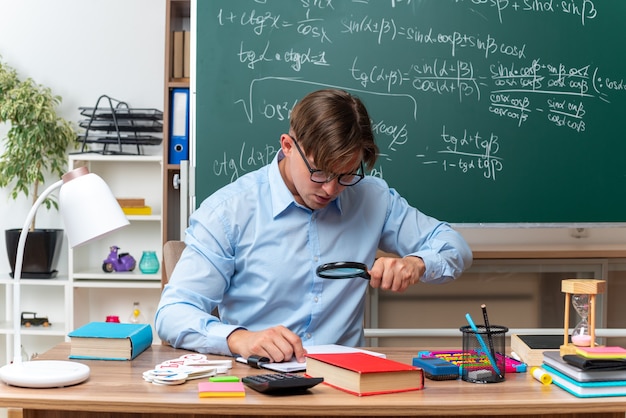 Giovane insegnante maschio con gli occhiali guardando le note attraverso la lente di ingrandimento preparando la lezione seduto al banco di scuola con libri e note davanti alla lavagna in aula