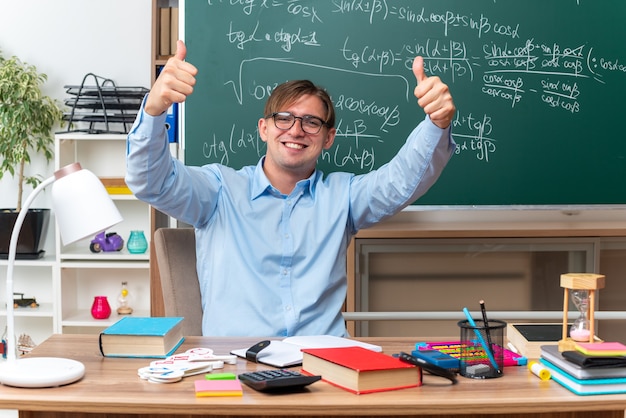 Young male teacher wearing glasses looking smiling cheerfully showing thumbs up sitting at school desk with books and notes in front of blackboard in classroom