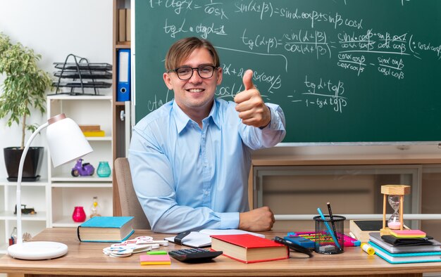 Young male teacher wearing glasses looking smiling confident showing thumbs up sitting at school desk with books and notes in front of blackboard in classroom