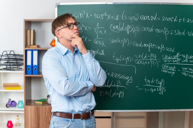 Young male teacher wearing glasses looking up puzzled standing near blackboard with mathematical formulas in classroom