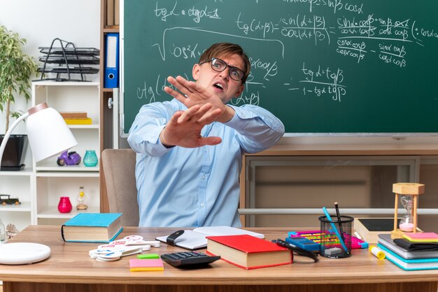 Young male teacher wearing glasses looking worried and confused making defense gesture with hands sitting at school desk with books and notes in front of blackboard in classroom