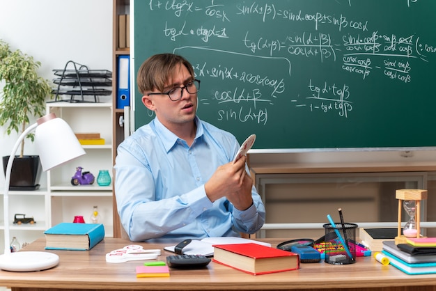 Young male teacher wearing glasses prepearing lesson looking confident sitting at school desk with books and notes in front of blackboard in classroom