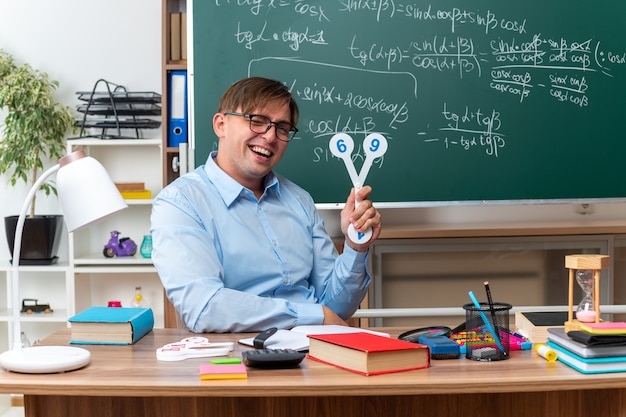 Молодой учитель-мужчина в очках показывает номерные знаки, объясняя урок, улыбаясь, сидя за школьной партой с книгами и заметками перед доской в классе