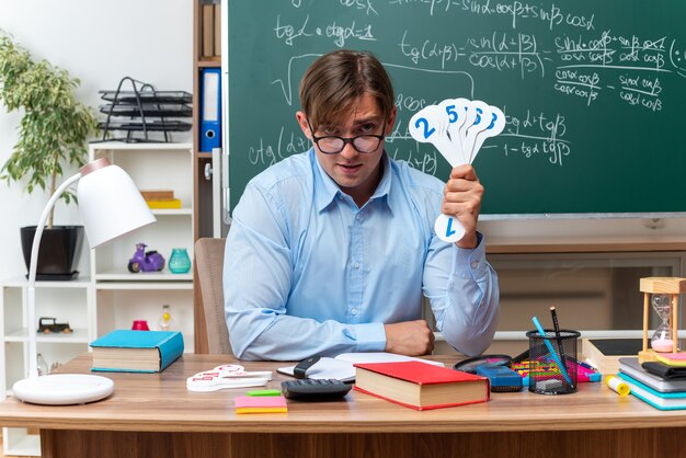 Young male teacher wearing glasses showing number plates explaining lesson smiling  sitting at school desk with books and notes in front of blackboard in classroom