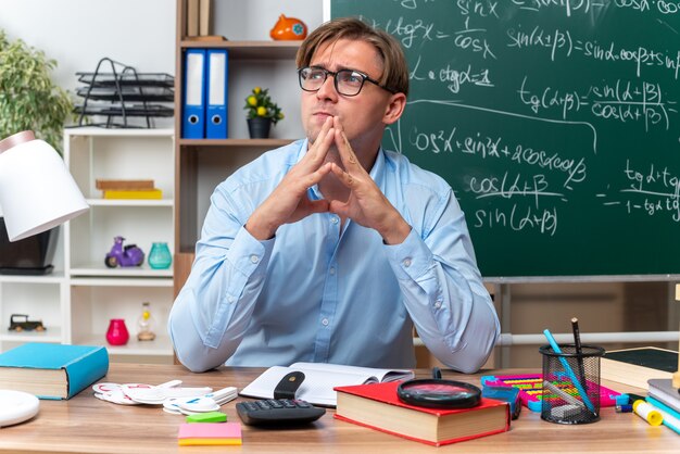 Young male teacher wearing glasses sitting at school desk with books and notes holding palms together looking aside with pensive expression on face thinking in front of blackboard in classroom