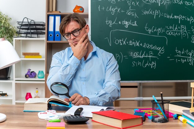 教室の黒板の前で真剣な顔で本を虫眼鏡で見る本とノートを持って学校の机に座っている眼鏡をかけた若い男性教師