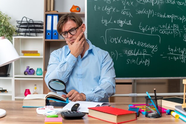 Young male teacher wearing glasses sitting at school desk with books and notes looking through magnifying glass at book with serious face in front of blackboard in classroom