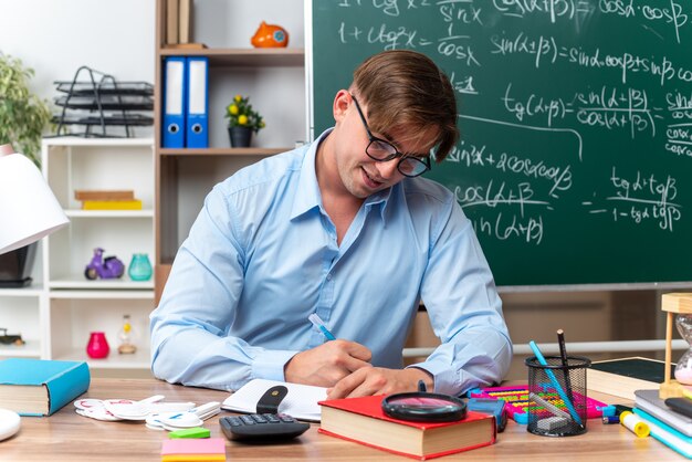 Young male teacher wearing glasses sitting at school desk with books and notes writing in front of blackboard in classroom