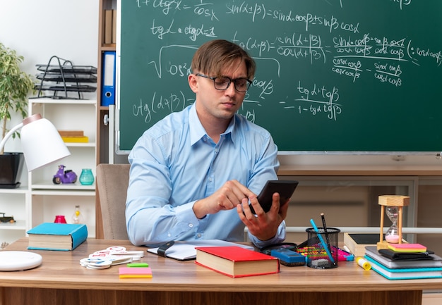 Young male teacher wearing glasses typing message using smartphone looking confident sitting at school desk with books and notes in front of blackboard in classroom