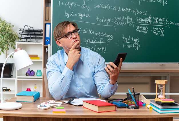 Молодой учитель-мужчина в очках печатает сообщение с помощью смартфона, выглядит озадаченным, сидя за школьной партой с книгами и заметками перед доской в классе