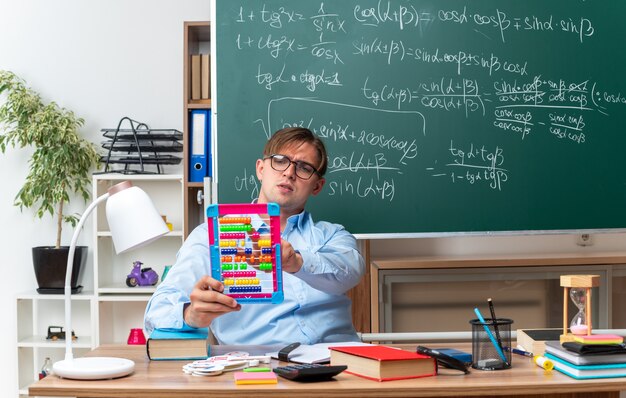 Young male teacher wearing glasses with bills looking confident preparing lesson sitting at school desk with books and notes in front of blackboard in classroom