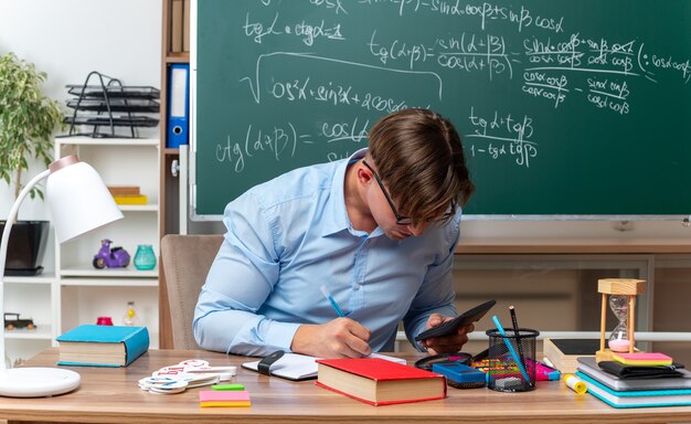 Молодой учитель-мужчина в очках с калькулятором готовит урок, выглядит уверенно, сидя за школьной партой с книгами и заметками перед доской в классе
