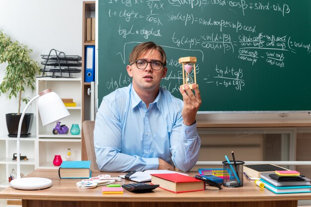 young male teacher wearing glasses with hourglass preparing lesson looking confident sitting at school desk with books and notes in front of blackboard in classroom
