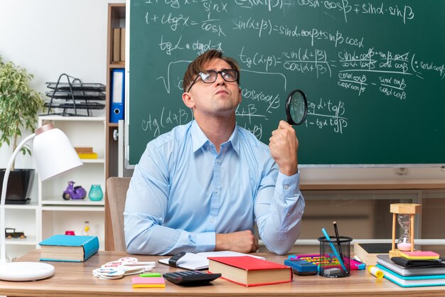 Young male teacher wearing glasses with magnifying glass preparing lesson looking up puzzled sitting at school desk with books and notes in front of blackboard in classroom