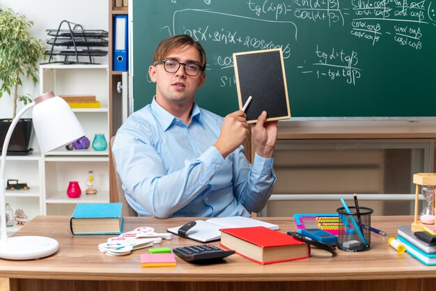 Young male teacher wearing glasses with small blackboard and piece of chalk explaining lesson looking confident sitting at school desk with books and notes in front of blackboard in classroom