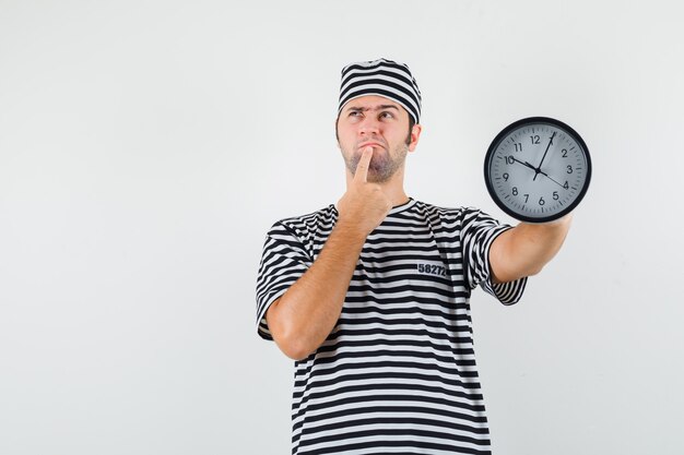 Young male thinking something while holding clock in striped t-shirt,cap and looking pensive , front view.