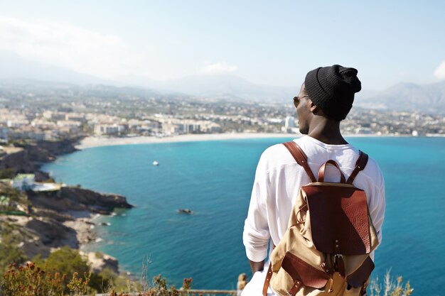 молодой путешественник с кожаным рюкзаком любуется бескрайним лазурным океаном и скалистым побережьем