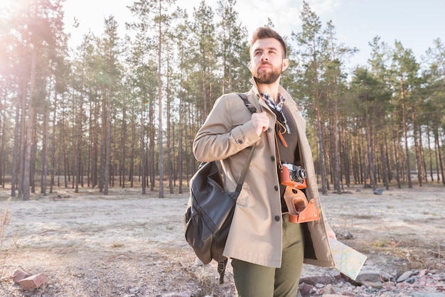 Young male traveler holding backpack on his shoulder and map standing in the forest