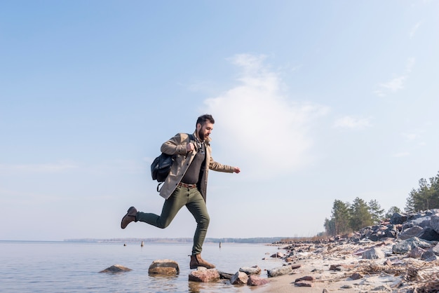湖の石の上を走っている彼のバックパックを持つ若い男性旅行者