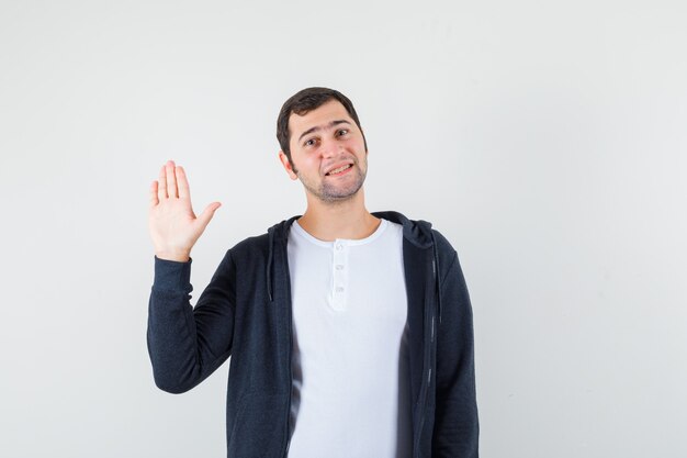Young male waving hand for greeting in t-shirt, jacket and looking cheery , front view.